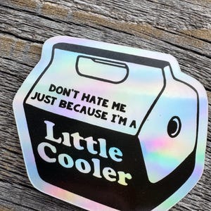 May include: A holographic sticker in the shape of a cooler. The sticker has the text "DON'T HATE ME JUST BECAUSE I'M A Little Cooler" in white and black. The sticker is on a wooden surface.