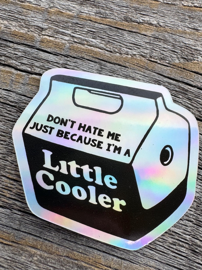 May include: A holographic sticker in the shape of a cooler. The sticker has the text "DON'T HATE ME JUST BECAUSE I'M A Little Cooler" in white and black. The sticker is on a wooden surface.
