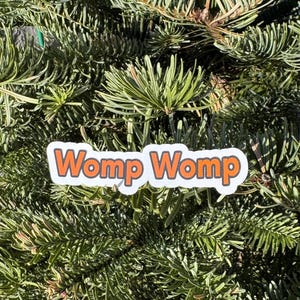 May include: A white sticker with orange text that reads "Womp Womp" on a green evergreen tree.