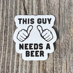 May include: White sticker with the text "THIS GUY NEEDS A BEER" in black, with thumbs-up hand illustrations. The sticker is on a weathered, gray wooden surface.