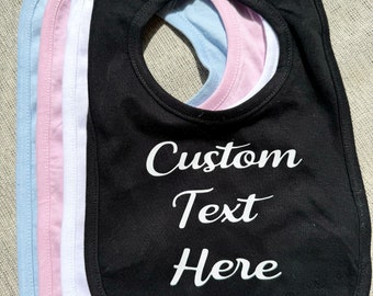 May include: A stack of four baby bibs in black, white, pink, and light blue. The top black bib features the white text Custom Text Here in a cursive font. Each bib has a rounded shape with a neck opening and a hook-and-loop closure at the top.