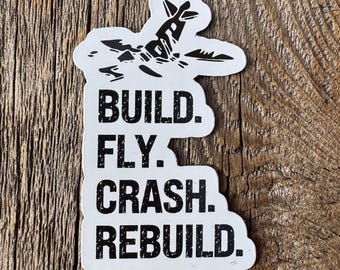 May include: A white sticker with black text and a graphic of an airplane. The text reads "BUILD. FLY. CRASH. REBUILD." The sticker is on a wooden surface.