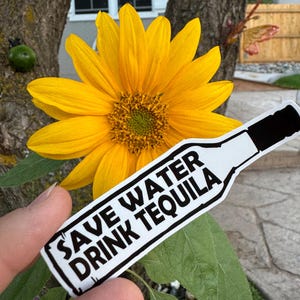 May include: A white sticker with black text that reads "Save Water Drink Tequila" and a silhouette of a bottle. The sticker is held in a hand in front of a yellow sunflower.