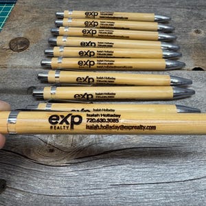 May include: A stack of wooden pens with a silver metal top and a black engraved logo that says "exp REALTY" and includes the name "Isaiah Holladay" and contact information.