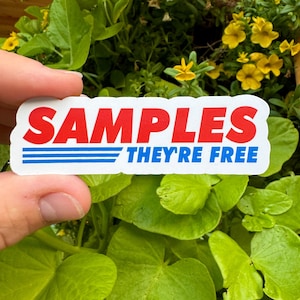 May include: A white sticker with blue and red text that reads "SAMPLES THEY'RE FREE". The sticker is held in a hand against a background of green leaves and yellow flowers.