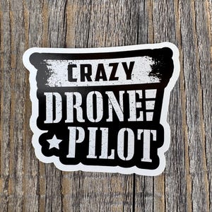 May include: A white sticker with black text that reads "CRAZY DRONE PILOT" with a star graphic. The sticker has a black border and is set against a weathered wood background.
