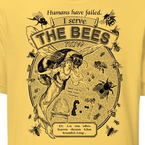 May include: A yellow t-shirt featuring a black and white illustration of a bee and a woman. The text on the shirt reads "Humans have failed. I serve THE BEES now. Bee town Thomas. Te is we who have done the bumbling."