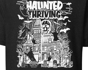 May include: Black t-shirt featuring a white graphic of a haunted house, ghosts, bats, and the words "Haunted and Thriving." The text "My demons are pretty cool, actually" is below the house.