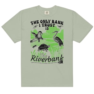 May include: Pale green t-shirt with a nature-themed graphic. The design includes a bird, heron, turtle, and dragonflies. Text reads "The Only Bank I Trust Is The Riverbank." Below, the text says "I gave all my money to a turtle! It just swam away."