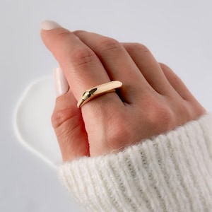 May include: A gold-toned ring with a thick, rectangular band. The ring is worn on the index finger of a hand.