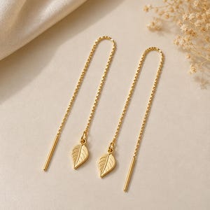 May include: A pair of gold-tone threader earrings featuring a delicate chain design. Each earring is finished with a small, polished metal leaf charm that hangs from the center of the chain, complemented by a straight metal bar at each end.