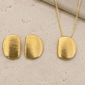 May include: A gold-toned jewelry set consisting of a necklace with a thin chain and a pendant, along with two matching stud earrings. Each piece features a semi-circular shape with a textured, brushed metal finish. The brand name NEBULAR JEWELS is visible.