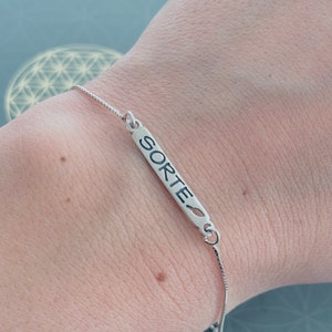 May include: A silver chain bracelet with a silver bar charm that says "SORTE" with a small leaf design.