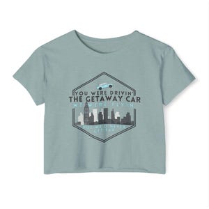 May include: A light blue cropped t-shirt with a graphic print of a car flying over a city skyline. The text on the graphic reads "You were drivin' the getaway car, we were flyin' but we'd never get far."