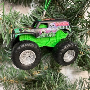 May include: A Grave Digger monster truck ornament with a green chassis and black tires. The truck has a black cab with the Grave Digger logo and a green suspension. A green string is attached for hanging on a Christmas tree.