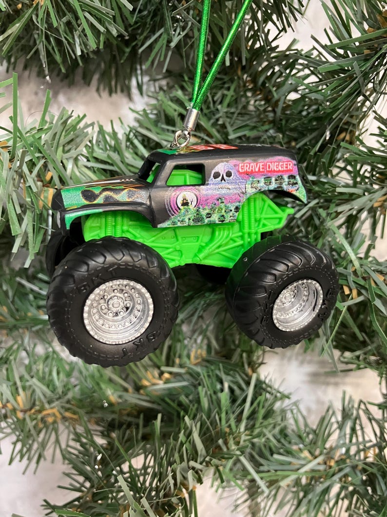 May include: A Grave Digger monster truck ornament with a green chassis and black tires. The truck has a black cab with the Grave Digger logo and a green suspension. A green string is attached for hanging on a Christmas tree.
