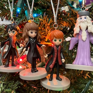 May include: A collection of Harry Potter character ornaments, including Harry, Hermione, Ron, and Dumbledore, hanging on a Christmas tree. Each figure is holding a wand and is on a circular base. The ornaments are colorful and detailed.