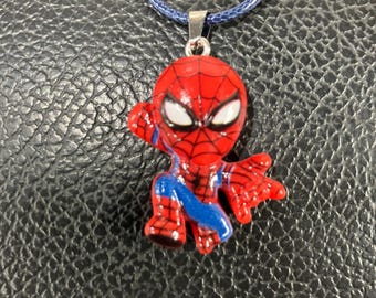 Pode incluir: Pingente do Homem-Aranha vermelho e azul em um colar de cordão azul. O pingente apresenta o design icônico da máscara e do traje do personagem. O colar é exibido em um fundo escuro.