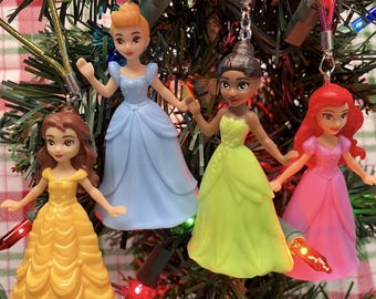 May include: Four princess-themed Christmas ornaments. Each doll is wearing a colorful gown: yellow, light blue, lime green, and pink. The ornaments are hanging on a Christmas tree with green needles and red and white plaid in the background.