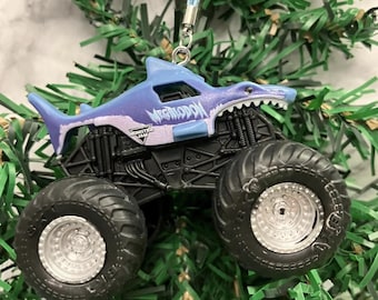 May include: A blue and black Monster Jam Megalodon truck ornament with a shark head design. The truck has large black wheels and the words "Monster Jam" and "Megalodon" are printed on the side.