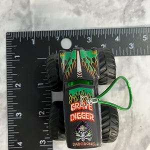May include: A black and green toy monster truck with the words "GRAVE DIGGER" and "BAD BONE" printed on the side. The truck has large black tires and a green loop attached. The truck is approximately 3 inches long.