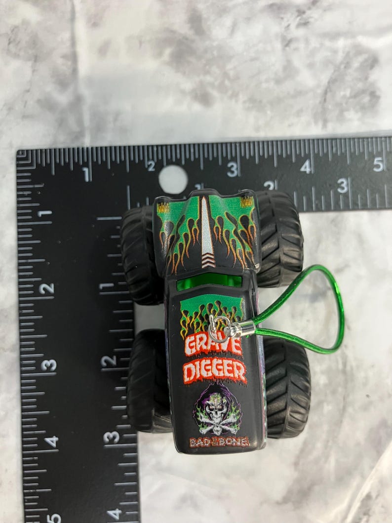 May include: A black and green toy monster truck with the words "GRAVE DIGGER" and "BAD BONE" printed on the side. The truck has large black tires and a green loop attached. The truck is approximately 3 inches long.