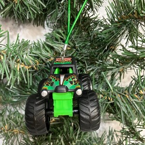May include: A black and green monster truck Christmas ornament with large black tires. The truck has a green front end and flames on the hood. A green string is attached for hanging on a Christmas tree.