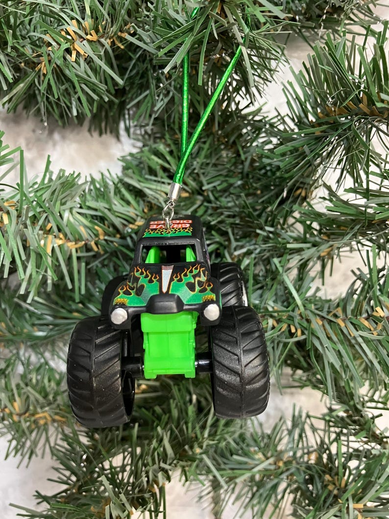 May include: A black and green monster truck Christmas ornament with large black tires. The truck has a green front end and flames on the hood. A green string is attached for hanging on a Christmas tree.