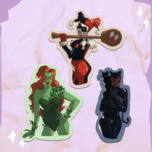 Op de afbeelding: Drie gestanste stickers met DC Comics-personages: Harley Quinn met een moker, Poison Ivy met groene huid en rood haar, en Catwoman met een zwarte kat. De stickers hebben een witte rand op een lichtpaarse achtergrond.
