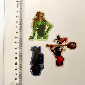 Op de afbeelding: Drie kleurrijke stickers met stripfiguren. E&eacute;n toont een vrouw met rood haar en groene klimop, een andere toont een vrouw in een zwart kattenpak, en de laatste toont een vrouw in een rood-zwart harlekijnkostuum.