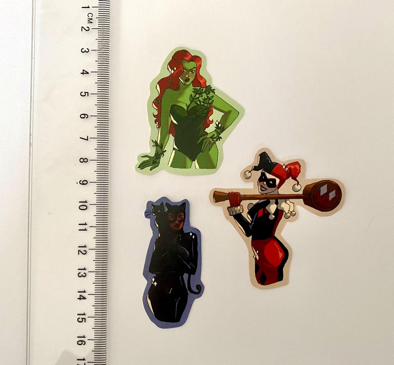 Op de afbeelding: Drie kleurrijke stickers met stripfiguren. E&eacute;n toont een vrouw met rood haar en groene klimop, een andere toont een vrouw in een zwart kattenpak, en de laatste toont een vrouw in een rood-zwart harlekijnkostuum.