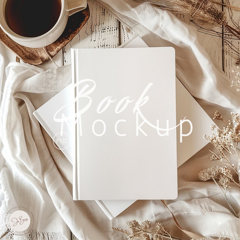 May include: A white book mockup with the text "Book Mockup" written in white on the cover. The book is lying on a white surface with a white fabric draped over it. There are dried flowers and a cup of coffee in the background.