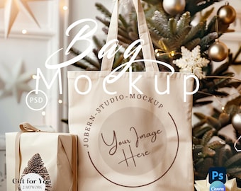 Puede incluir: Una maqueta de bolsa de tela beige con el texto "Your Image Here" en un círculo en la parte delantera. La bolsa está sobre una superficie blanca con una caja de regalo marrón y un árbol de Navidad en el fondo.