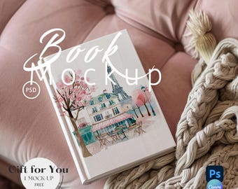 Puede incluir: Una maqueta de libro blanca con una ilustración de acuarela de una escena de café parisino con un edificio rosa, un toldo rosa, un árbol con flores rosas y la Torre Eiffel. El libro está sobre un sofá de terciopelo rosa con una manta de punto blanco.