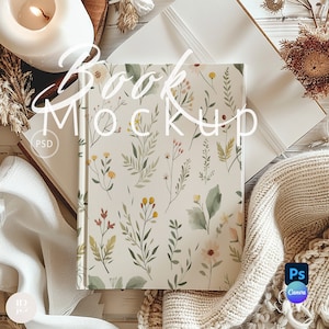 May include: A book mockup featuring a floral design on the cover. The book is surrounded by a white blanket, a candle, and dried flowers. The text "Book Mockup" is written in white on the image.