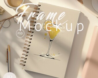 May include: A white notebook with a gold spiral binding. The notebook features a drawing of a cocktail glass with a lemon wedge on the rim. The text "Frame Mockup" is written in black cursive on the notebook. The notebook is on a white surface with a gold pen, a pair of glasses, and a cup of tea.