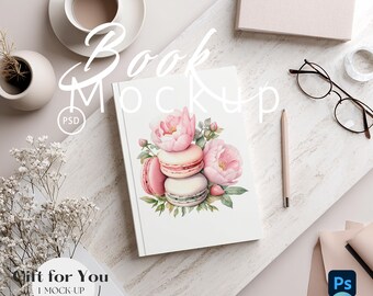 May include: A white book mockup with a watercolor design of pink macarons and flowers. The image includes a cup of coffee, glasses, a pencil, and other stationery items. Text on the book cover reads "Book Mockup".