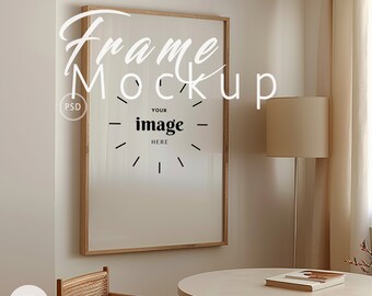 May include: A wooden frame mockup with a white background and the text "Your image here" inside the frame. The frame is hanging on a light beige wall. A small table with a book on it is in the foreground.