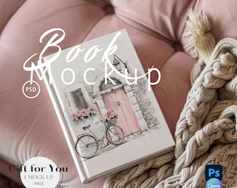 May include: A white book mockup with a watercolor illustration of a pink door, flowers, and a bicycle on a brick building. The book is lying on a pink velvet couch with a cream knitted blanket.