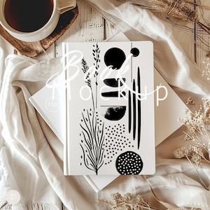 May include: A white notebook with a black and white abstract design featuring a plant, circles, and lines. The notebook is on a white surface with a white cloth and dried flowers.