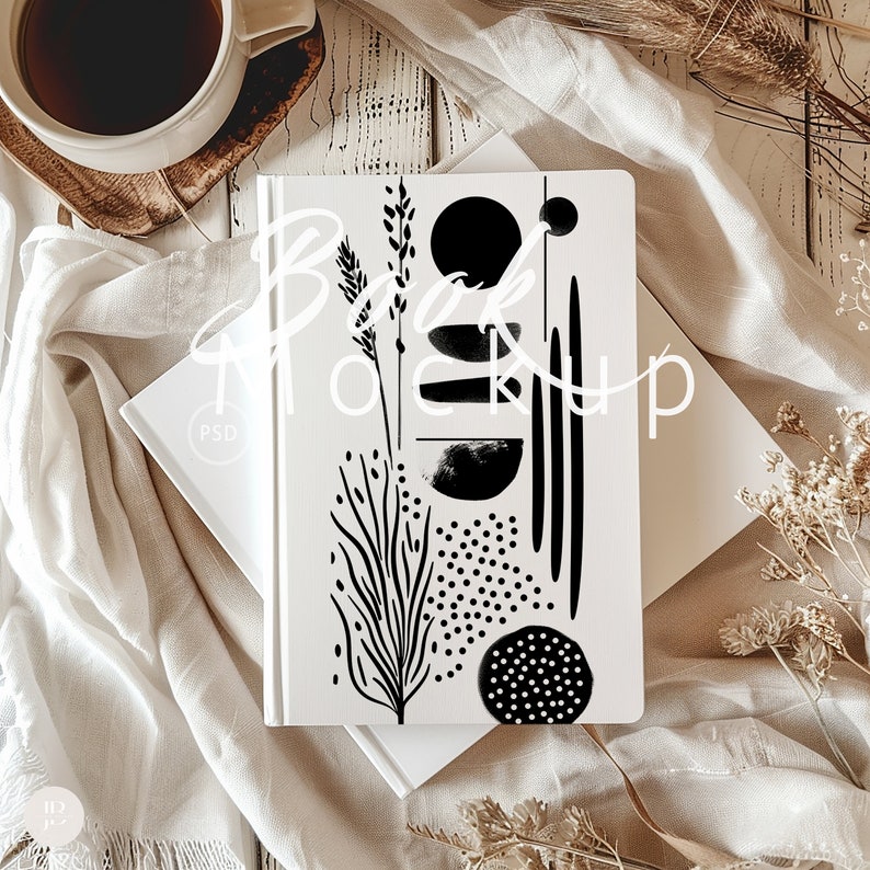 May include: A white notebook with a black and white abstract design featuring a plant, circles, and lines. The notebook is on a white surface with a white cloth and dried flowers.