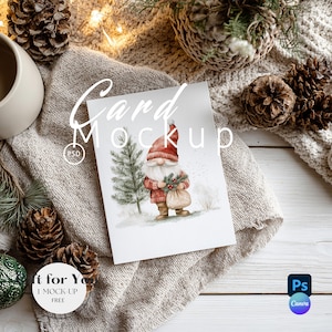 May include: A greeting card mockup featuring a watercolor illustration of a gnome holding a sack and greenery, with a Christmas tree. The card is on a neutral-toned fabric, surrounded by pine cones and a mug. The words "Card Mockup" are written on the card.