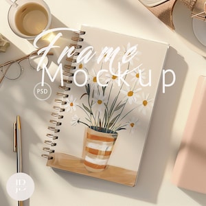 May include: A white notebook with a spiral binding and a floral illustration of white daisies in a striped orange pot. The notebook has the text "Frame Mockup" written in black cursive on the front cover. The notebook is on a white surface with a gold pen and a pair of glasses.