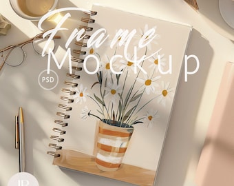 May include: A white notebook with a spiral binding and a floral illustration of white daisies in a striped orange pot. The notebook has the text "Frame Mockup" written in black cursive on the front cover. The notebook is on a white surface with a gold pen and a pair of glasses.