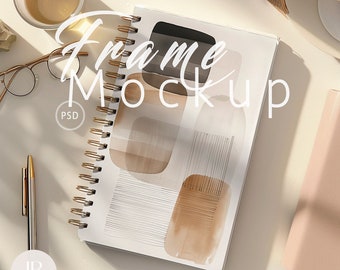 May include: A white spiral bound notebook with a brown and white abstract design. The notebook is on a white surface with a gold pen, a pair of glasses, and a cup of tea.