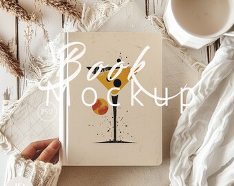 May include: A book mockup with a beige cover and a stylized illustration of a martini glass with a black dot and a yellow circle. The text "Book Mockup" is written in white cursive on the cover.
