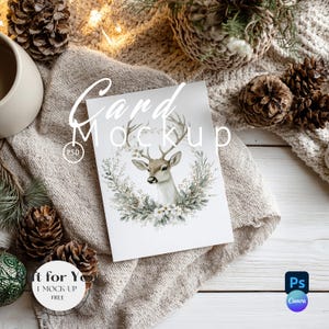 Op de afbeelding: Een mockup van een wenskaart met een aquarel illustratie van een hertenkop met geweien, omringd door een bloemenkrans. De kaart ligt op een neutrale stof met dennenappels en een mok. De tekst "Card Mockup" staat op de kaart.