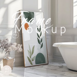 May include: A framed art print with a gold frame, featuring abstract botanical shapes in green, orange, and white. The artwork leans against a white wall in a bathroom setting. The text "Frame Mockup" is overlaid on the image.