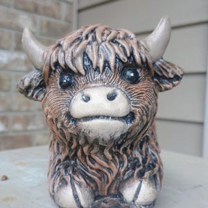 May include: A ceramic Highland cow figurine with a brown and tan color scheme. The cow has large, dark eyes, small horns, and a shaggy coat. The figurine is sitting on a gray surface.