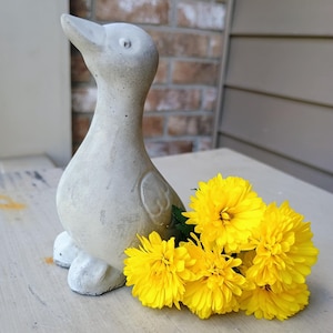 May include: A concrete duck ornament with a weathered, textured finish. The duck is light grey and has a long neck and a slightly open beak. Bright yellow flowers are arranged beside the duck, providing a vibrant contrast.